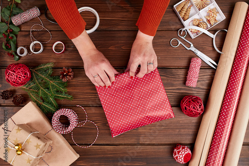 Woman s hands wrapping Christmas gift, close up. Unprepared christmas presents on wooden background with decor elements and items, top view. Christmas or New year DIY packing Concept. photo