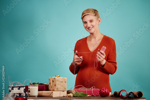 Pretty blond young mother packing Christmas presents for her children holding scissors and backer s twine stripe in hands, isolated at studio over blue background. Present packaging ideas. photo