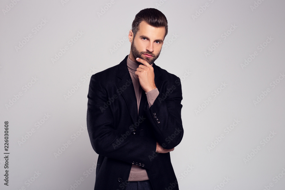 Hmmmmmm. Handsome young man in coat looking at camera and keeping hand on chin while standing against white background Hmmmmmm. Handsome young man in coat looking at camera and keeping hand on chin while standing against white background