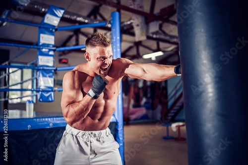 Male boxer training with punching bag in dark sports hall. Young boxer training on punching bag. Male boxer as exercise for the big fight. Boxer hits punching bag.