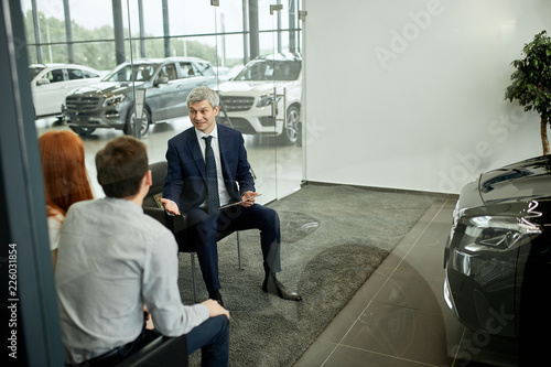 Caucasian couple is buying new car, mature seller in a formal wear is presenting contract to sign, in his office at car dealership