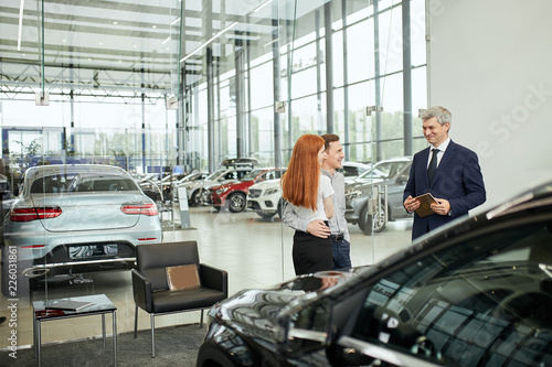 Sales agent at the table desk telling about car features to cheerful couple of customers at car dealership office