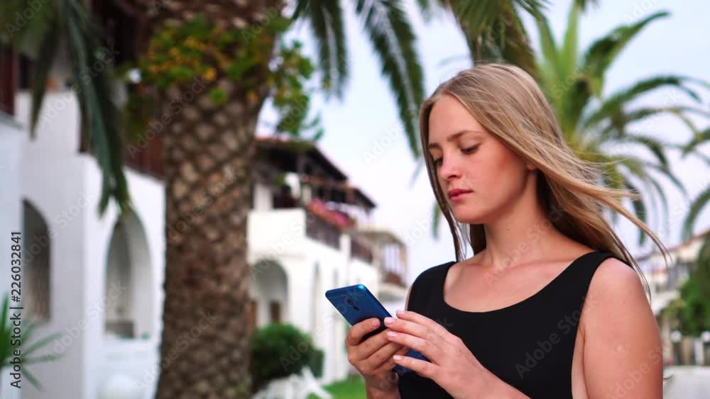 Busy relaxed dreamy young girl looking at smart phone device screen reading in middle of palm tree tropical resort alley Busy relaxed dreamy young girl looking at smart phone device screen reading in middle of palm tree tropical resort alley