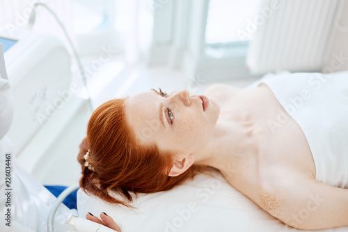 Attractive red haired woman lying on couch in medical centre, waiting for beauty massage. Facial treatment. Cosmetology, beauty and skin care and treatment concept .