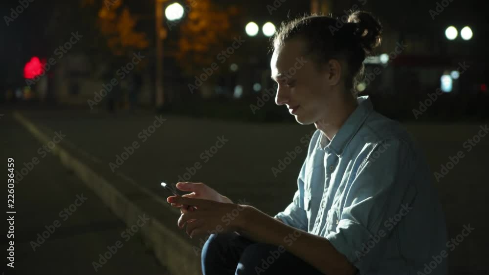  Profile of a stylish young man with a bun haircut browsing the net cheery on his smartphone and seeking info in a green park deep at night in autumn