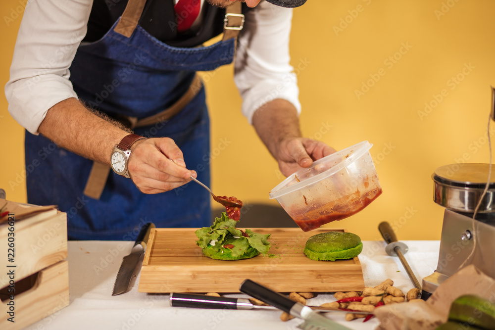 Caucasian bearded man in apron and cap making hamburger, adding ingredients on green bun, isolated studio shot on yellow background