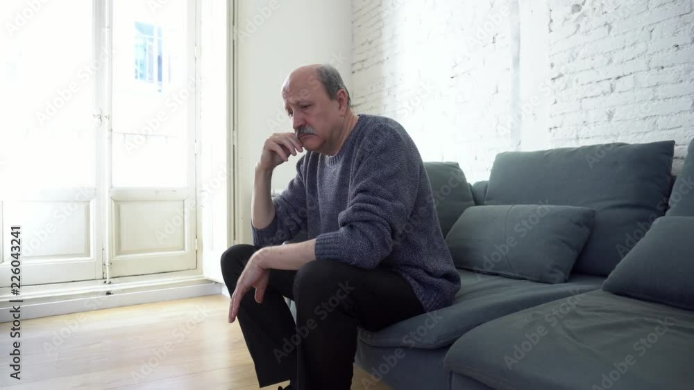 Thoughtful depressed and lonely senior old man sitting on his couch at home looking out the window in depression mental health concept. Thoughtful depressed and lonely senior old man sitting on his couch at home looking out the window in depression mental health concept.