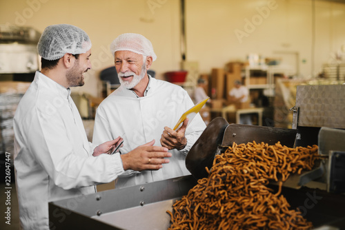 Proud father and son in sterile clothes standing in their food factory and checking the quality of products. Smiling and hands gesturing to each other.
