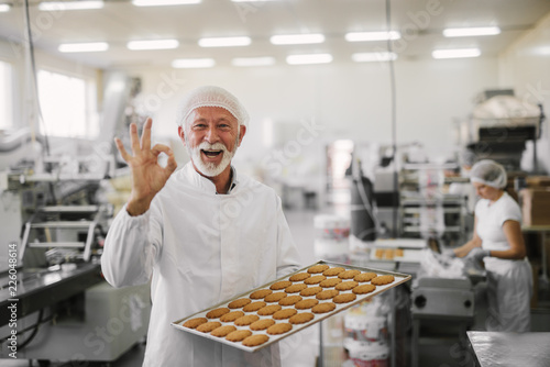 Good quality.Picture of mature cheerful male employee in sterile clothes standing in bright food factory . Holding tray full with fresh cookies and gesturing with his hand that quality is good.