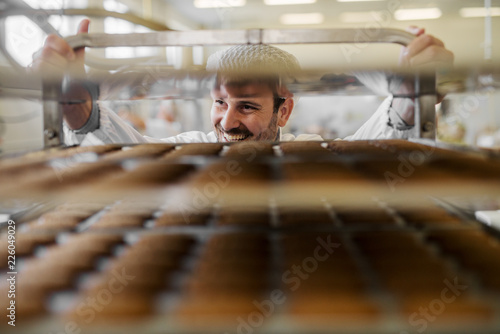 Picture of professional male baker man in white work uniform . Standing in front of the shelves full with fresh baked cookies.