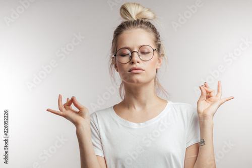 Concentrated beautiful young woman with blonde hair knot dressed in white t-shirt keeping eyes closed while meditating indoors, practicing peace of mind, keeping fingers in mudra gesture photo