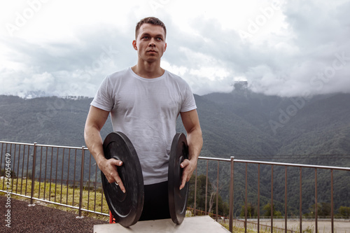 Outdoor shot of sportsman in in training wear, standing on mountain top with two barbell weight discs over mountain forest and sky background. photo