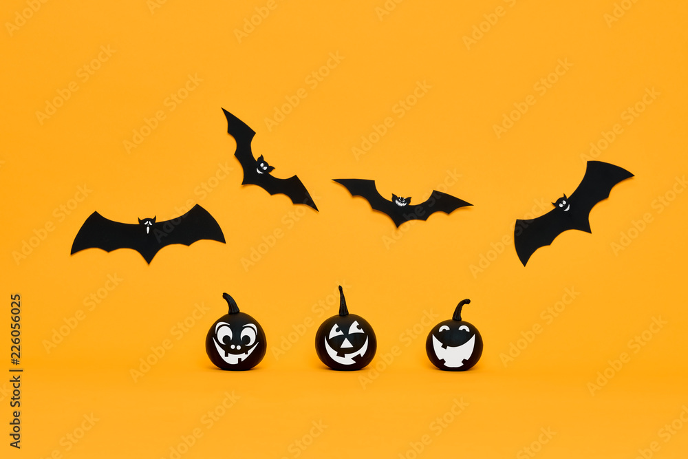 Cute Halloween pumpkins with funny smiling faces and paper bats flying over orange background. Halloween concept.