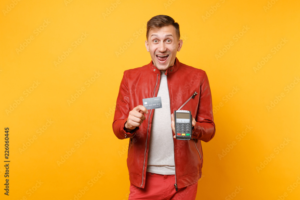 Portrait man in red leather jacket, t-shirt hold wireless modern bank payment terminal to process acquire credit card payments isolated on yellow background. People lifestyle concept. Advertising area Portrait man in red leather jacket, t-shirt hold wireless modern bank payment terminal to process acquire credit card payments isolated on yellow background. People lifestyle concept. Advertising area