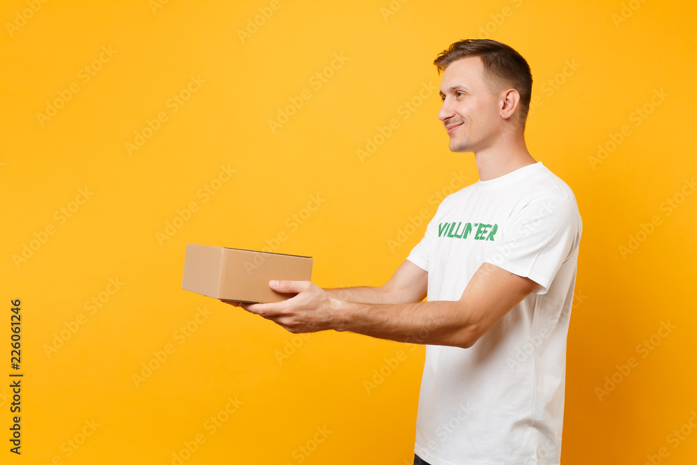 Side profile view of man in white t-shirt with written inscription green title volunteer with blank cardboard box isolated on yellow background. Voluntary free assistance help, charity grace concept. Side profile view of man in white t-shirt with written inscription green title volunteer with blank cardboard box isolated on yellow background. Voluntary free assistance help, charity grace concept.