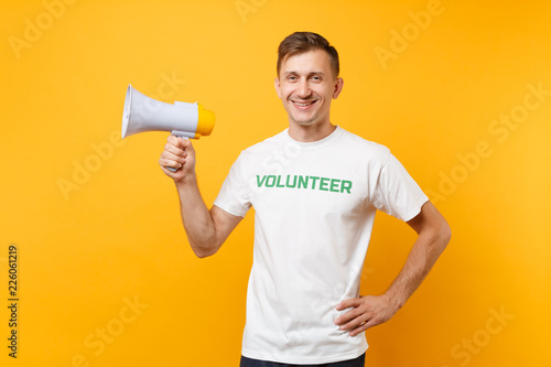 Portrait of man in white t-shirt written inscription green title volunteer scream in public address megaphone isolated on yellow background. Voluntary free assistance help, charity grace work concept.