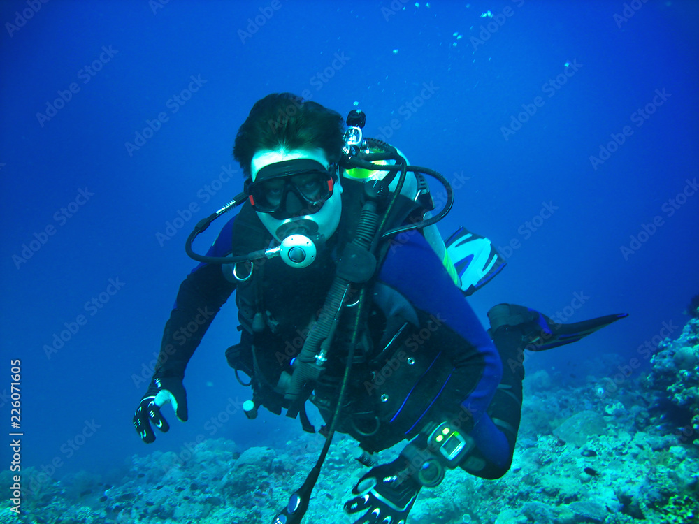 Scuba diver is underwater. He is wearing in full scuba-diving equipment: mask, regulator, BCD, fins. Diver is on the blue water background and corals are on the bottom. Scuba diver is underwater. He is wearing in full scuba-diving equipment: mask, regulator, BCD, fins. Diver is on the blue water background and corals are on the bottom.