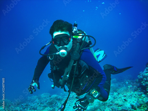 Scuba diver is underwater. He is wearing in full scuba-diving equipment: mask, regulator, BCD, fins. Diver is on the blue water background and corals are on the bottom.