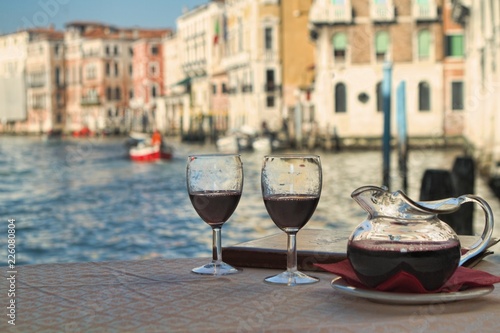 A carafe and two wine glasses filled with red wine sit on a table in front of a leather-bound menu at a restaraunt overlooking the Grand Canal in Venice, Italy