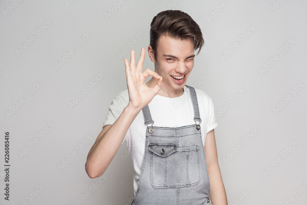 Happy young brunette man in casual style with t-shirt and denim overalls standing and looking at camera with toothy smile, winking and showing Ok sign. indoor studio shot, isolated on gray background
