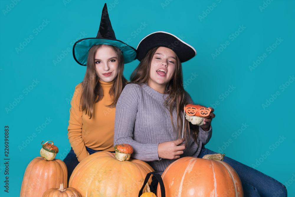 Fototapeta premium Portrait of two happy young women in black witch halloween costumes on party over blue background. The concept of Halloween, funny faces. Teenage girls with pumpkin and attributes of the holiday.