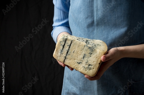 Woman in apron holding piece of blue cheese Gorgonzola on dark background photo