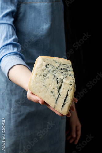 Woman in apron holding piece of blue cheese Gorgonzola on dark background