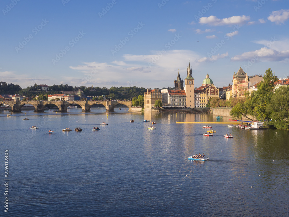 panorama of Charles bridge over Vltava river and houses of Old Town, Prague, Czech Republic, golden hour light, summer sunny day, tourists relaxing on pedal boats