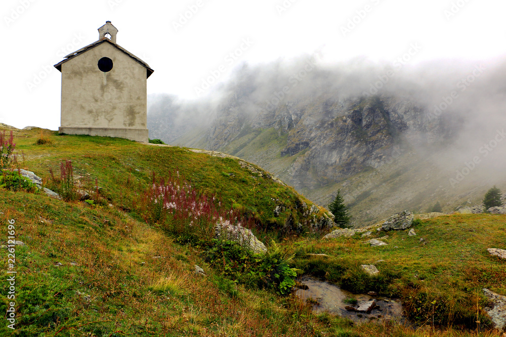 Back of small church beside brook on the way to Monviso, in Itaian Alps on the top of on one hill with dense fog behind and very high mauntain