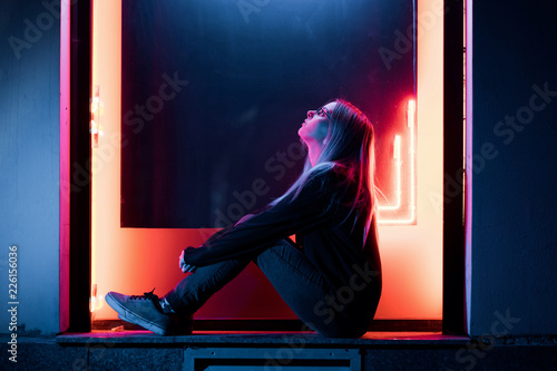 Millennial enigmatic pretty girl with unusual dyed hairstyle near glowing neon wall at night. Blue hair, golden sequins as freckles,nose piercing. Mysterious hipster teenager in glasses photo