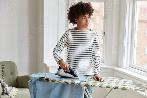 Valokuva Thoughtful dark skinned young housewife in striped clothes irons clothes on ironing board, uses electric iron stands in laundry, looks out of window, being busy with housework