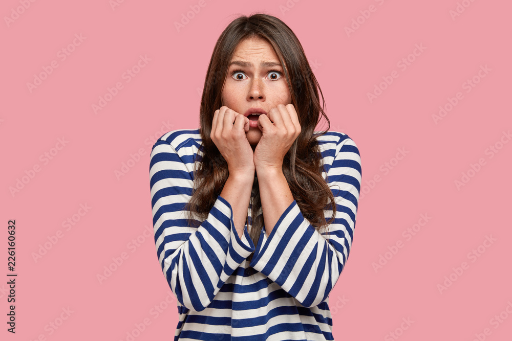 Scared European girl stares at camera with unexpected expression, dressed in sailor striped sweater, keeps hands near mouth, isolated over pink background. Young woman notices something scarying Scared European girl stares at camera with unexpected expression, dressed in sailor striped sweater, keeps hands near mouth, isolated over pink background. Young woman notices something scarying