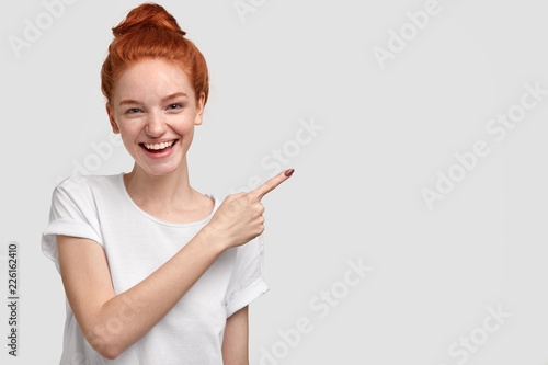 Glad satisfied red haired young female with charming smile, shows free space for your promotional content, advertises something against white background, dressed casually. Look aside, please.