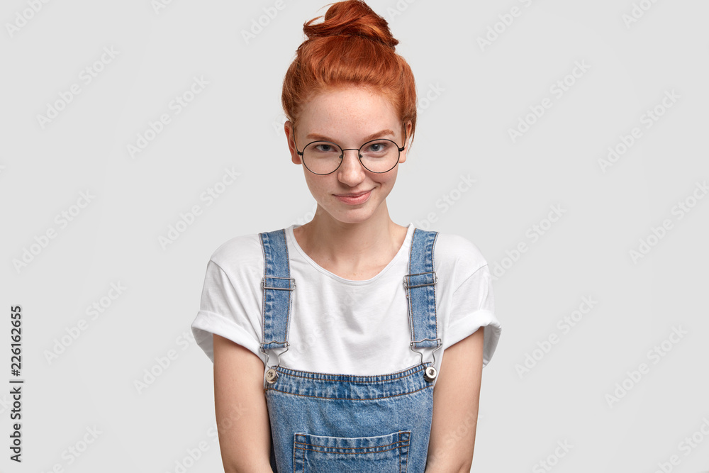 Φωτογραφία Horizontal shot of pleasant looking ginger young European woman in round transparent glasses, stylish dungarees, poses against white background, has glad expression