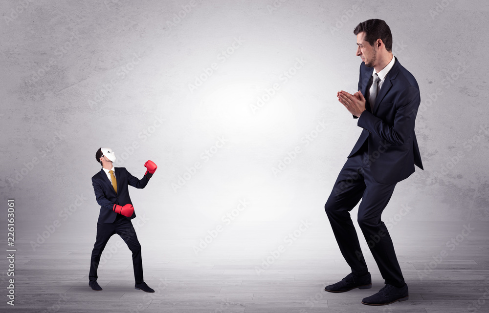 Big businessman being afraid of small masked businessman with box gloves in an empty room concept
Big businessman being afraid of small masked businessman with box gloves in an empty room concept