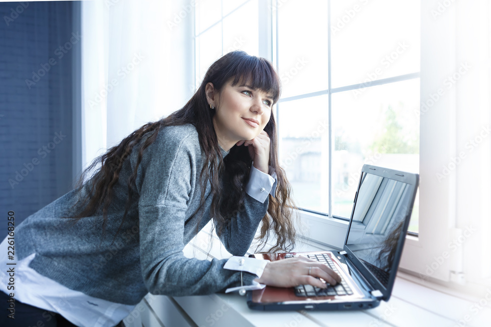 Middle-aged pretty woman is working with the PC notebook at home near the window Middle-aged pretty woman is working with the PC notebook at home near the window