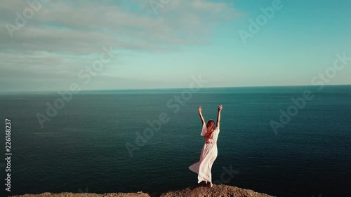 From the back. The brunette in pink dress is standing on a cliff near the ocean with hands up and looking at the beautiful horizon with blue sky and turquoise ocean in sunset light. Gran Canaria. photo