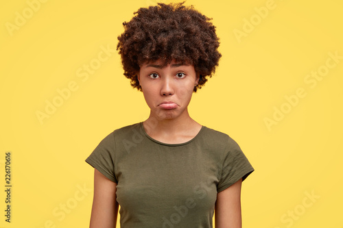 Photo of offended dark skinned girl being insulted by close person, dressed in casual militayr t shirt, expresses negative emotions, poses against yellow background. Why have you eatten all cookies? © wayhome.studio 