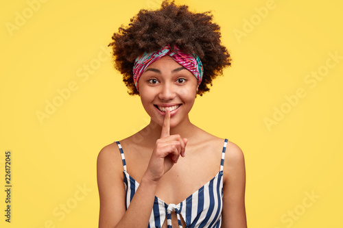 Beautiful dark skinned cheerful model wears casual striped top, colourful headband on forehead, keeps index finger over mouth, asks not spread rumors, isolated over studio yellow background.