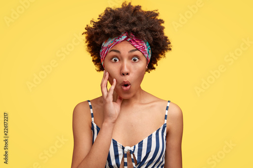 Cannot believe its true. Stupefied beautiful black impressed housewife  keeps mouth opened  holds palm on cheek  stares with scared expression  poses against yellow background. Reaction concept