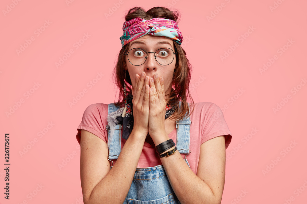 Horizontal View Of Surprised Emotive Female Hippy Keeps Both Hands On Mouth Has Bugged Eyes Cant Believe In Stunning News Wears Denim Overalls Poses Against Pink Background Reaction Concept Stock Photo