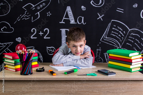 Schoolboy sitting at the desk with a chin leaned on the hands, surrounded with school supplies. Chalkboard as a background