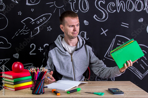 Contempt youngster sitting at the desk with a pile of books in the hand, surrounded with school supplies. Chalkboard as a background