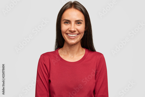 Photo of satisfied brunette woman with dark hair, has pleasant smile, being in good mood, wears red casual sweater, isolated over white background. Tanned lady rejoices positive moment in life photo