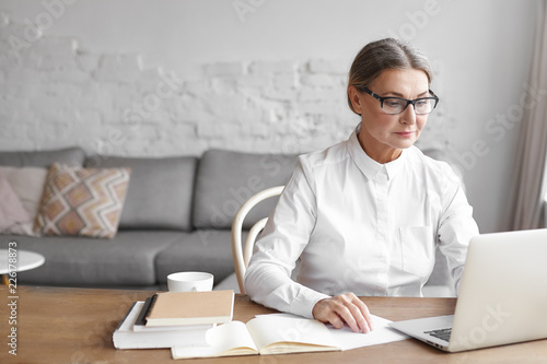 Busy modern mature female wearing spectacle and formal shirt working distantly using free wifi on generic laptop, sitting at wooden desk with books, having focused concentrated facial expression