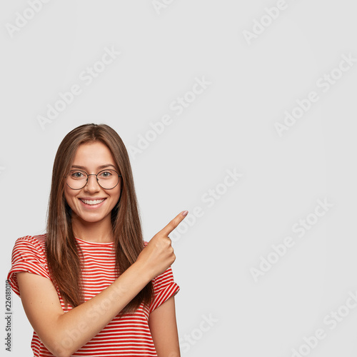 Indoor shot of young European cute woman with dark straight hair, charming smile, points aside with index finger, shows free space for your advertisement, wears casual t shirt, models indoor.
