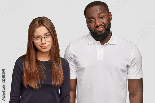 Two multiehnic teenage friends squint and frown, have displeased facial expressions, gaze with careless looks, being fed up with uninterested story of interlocutor, stand next to each other indoor