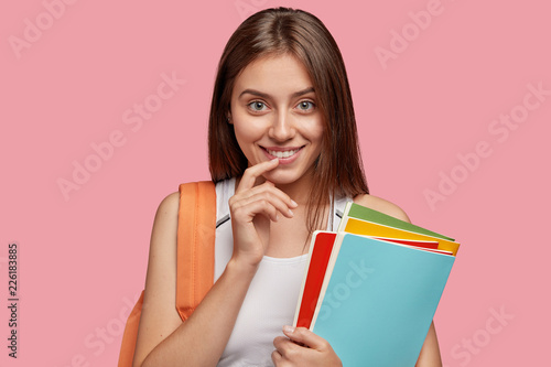 Good looking brunette woman with satisfied expression, keeps index finger near mouth, smiles gently, has intrigued look, thinks about creative plan, studies at college or university, uses books