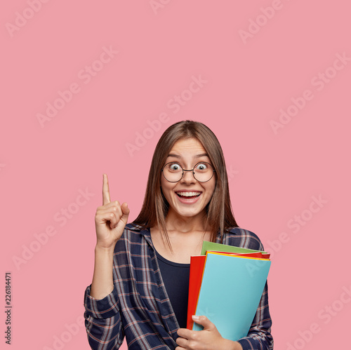 Photo of surprised European woman points with index finger upwards, being in good mood, advertises something upwards, dressed in checkered shirt, carries textbooks, stands against pink background