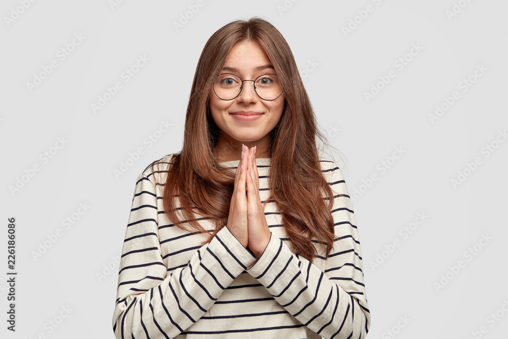 Pleased attractive woman with gentle smile, keeps hands in pray, dressed in striped clothes, asks for one more apology, has pleading expression, models against white background. Give me favour Pleased attractive woman with gentle smile, keeps hands in pray, dressed in striped clothes, asks for one more apology, has pleading expression, models against white background. Give me favour